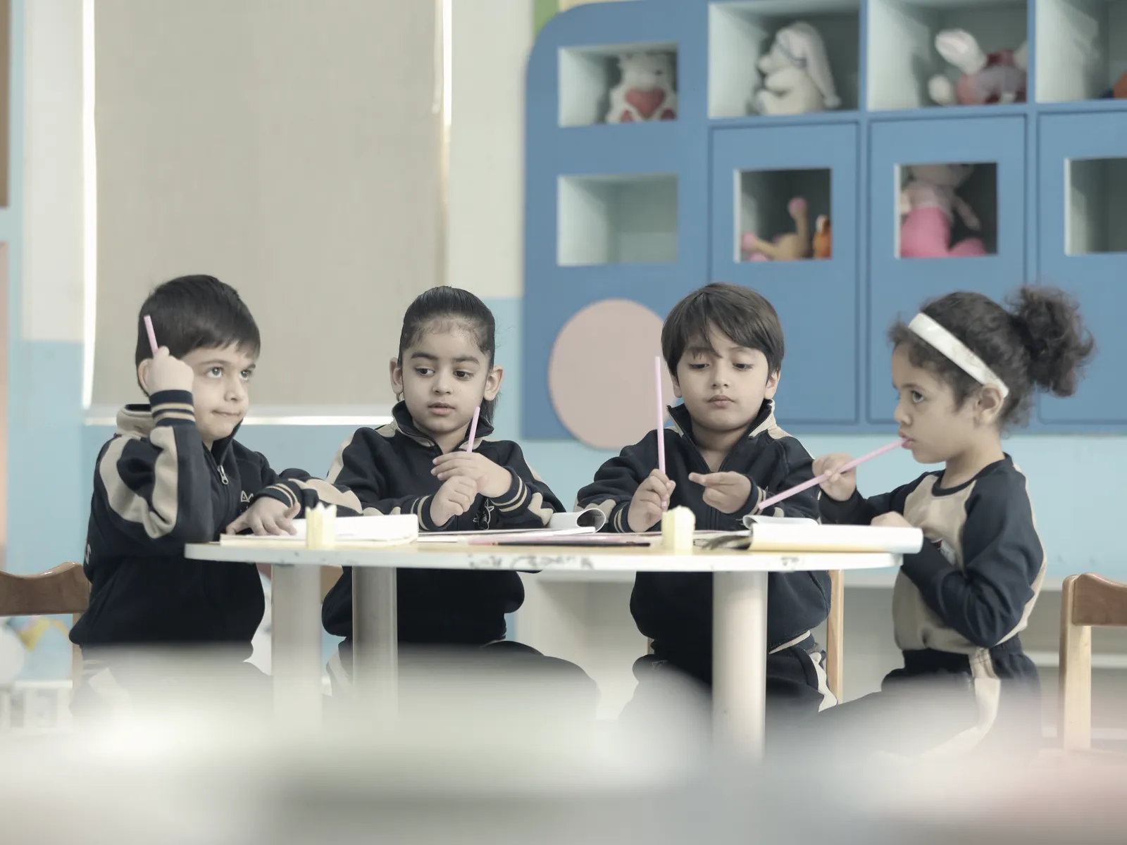 Four learners at a low round table in a Small Wonders classroom, drawing, with soft toys on the shelves behind them.